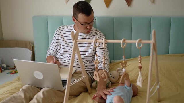 Young multitasking father trying to work on his laptop from the comfort of his bed while caring for his infant son, illustrating the challenges of parental leave and remote work
