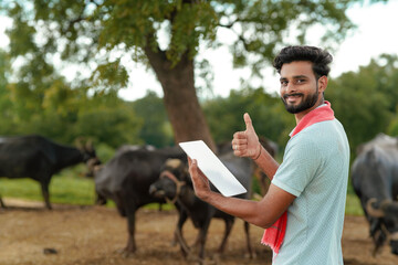 Fototapeta premium A young Indian farmer standing outdoors with his livestock holding a pad and giving a thumbs-up gesture.