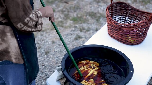 Retired craftswoman mixing fabric inside dye bath outdoors, handmade textile production, practical craft skill, self employed business, active retirement, productive aging lifestyle.