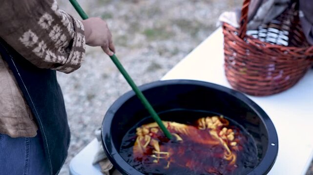 Active senior craftswoman working over dye bath outdoors, tie dye textile process, handmade production, small business routine, craft expertise, independent retirement lifestyle work.