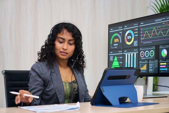 Professional business woman with curly hair wearing headset working on data analytics dashboard in modern office using tablet and financial documents for business strategy and customer support.