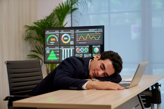 Exhausted businessman in suit sleeping at office desk with laptop and large data analytics dashboard in background, representing burnout, overwork, and stressful corporate work environment.
