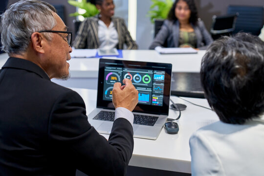 Senior male executive presenting data analytics dashboard on laptop during business meeting in corporate boardroom with diverse team, focus on financial growth, strategy, and leadership.