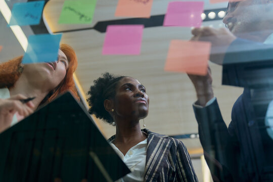 Diverse team of creative professionals brainstorming on glass wall with colorful sticky notes in modern office during strategy meeting and workshop for startup innovation project.