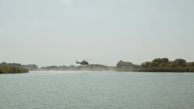 Wide shot of an Airbus H215 Super Puma firefighting helicopter refilling its Bambi bucket on a lake and flying away in Deliblatska Pescara. Deliblato, Serbia - August 7, 2026.
