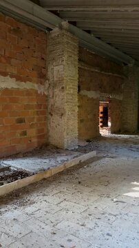 Vertical slow motion shot of the interior of an abandoned Spanish country house attic. The scene shows a mix of older brickwork, concrete beams, newer hollow brick partitions, and dust on the floor un