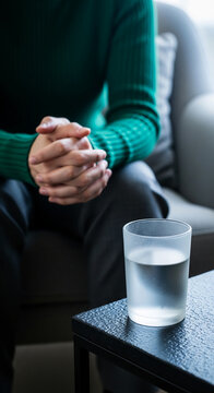 Person sitting with clasped hands next to a glass of water, symbolizing contemplation, patience, or waiting, suggestive of emotional state or therapy.