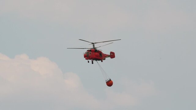Tracking shot of a red Kamov Ka-32 firefighting helicopter flying with a Bambi bucket through smoke during wildfire suppression operation. Deliblato, Serbia - August 7, 2026.