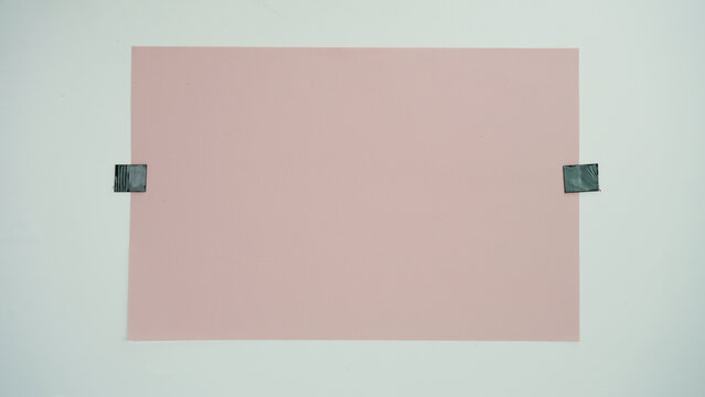 A piece of pink paper positioned horizontal and attached to a surface with black tape on two sides, isolated on a white background.