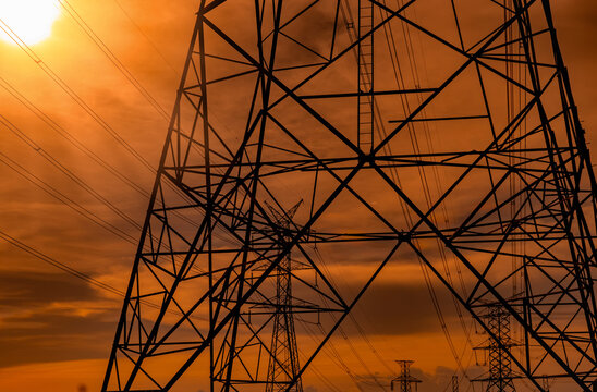 High voltage electricity transmission towers silhouetted against dramatic orange sunset sky. Modern power grid infrastructure for energy distribution, utility engineering and electrical network.