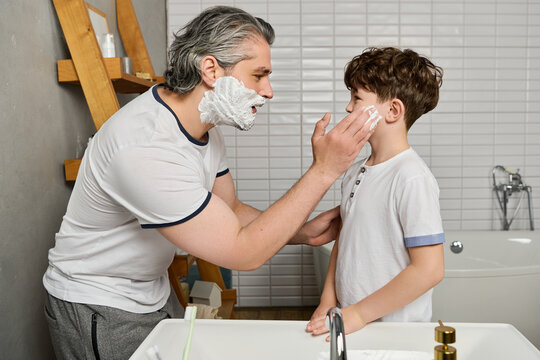 A dad playfully puts shaving cream on his sons face in the bathroom, making a fun memory.