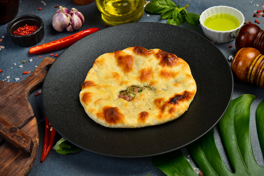 Freshly baked meat flatbread served on round dark plate. Traditional Georgian kubdari khachapuri pie placed on table. Tasty homemade pastry surrounded by spices and chili.
