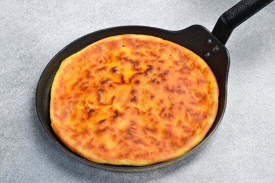 Traditional roasted round pie baked inside black frying pan top view. Tasty golden cheese flatbread surrounded by garlic and chili peppers. Gourmet homemade meal recipe.