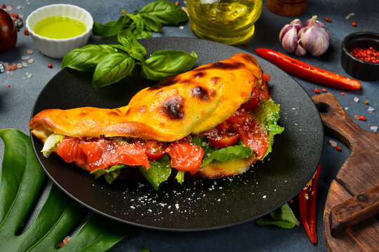 Tasty Italian folded pizza stuffed with smoked salmon. Fresh lettuce leaves and ripe tomatoes fill the baked dough. Delicious gourmet lunch served on dark dish.