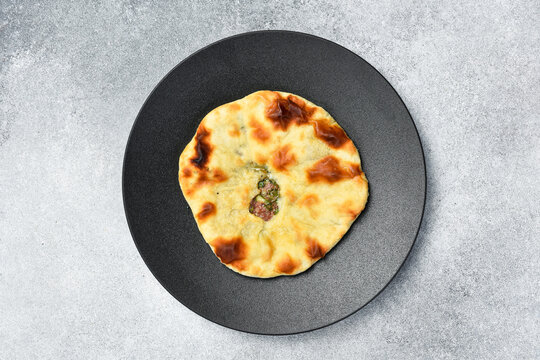 Freshly baked meat flatbread served on round dark plate. Traditional Georgian kubdari khachapuri pie placed on table. Tasty homemade pastry surrounded by spices and chili.