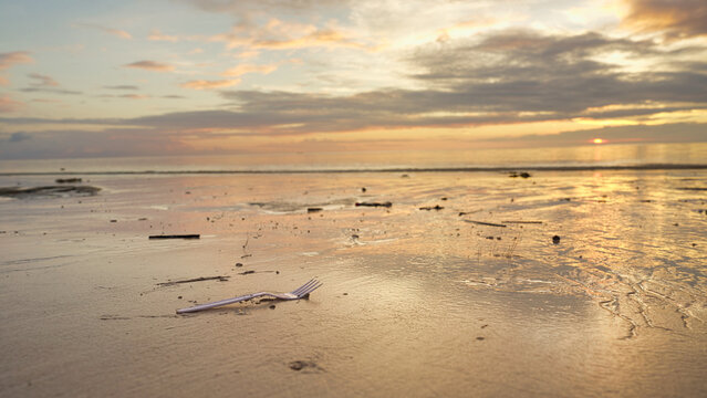 single-use plastic fork on sand at beach, ocean pollution, sunset and clouds, sea water