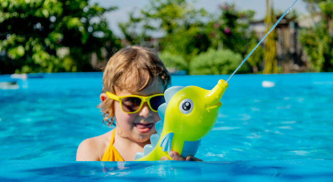 Little girl in sunglasses shooting water gun in swimming pool, water jet stream.