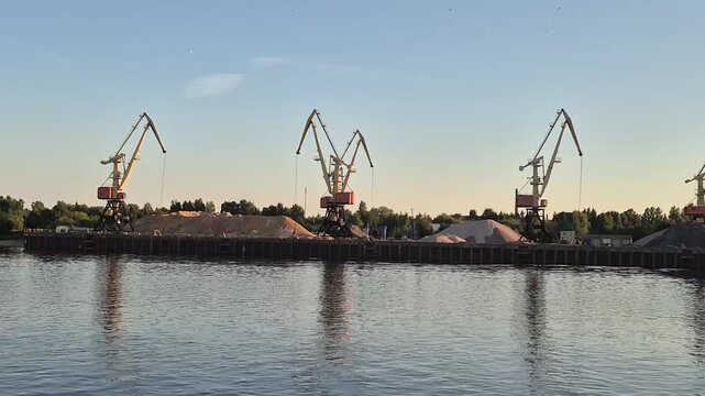 Industrial port cranes at river bank during summer trip. View of sand piles and cargo machinery from water transport. River travel landscape and busy dockyard.