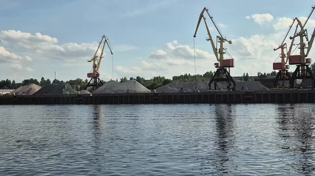 View of river port with industrial cranes and stone piles from tourist ship. Cargo terminal during summer journey along waterway. Sightseeing of port facility.