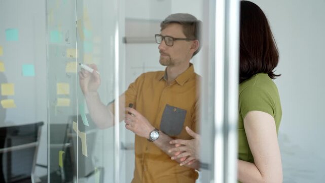 Business colleagues collaborate at a modern office glass board, mapping a timeline with sticky notes and hand drawn charts during an engaged strategic planning session