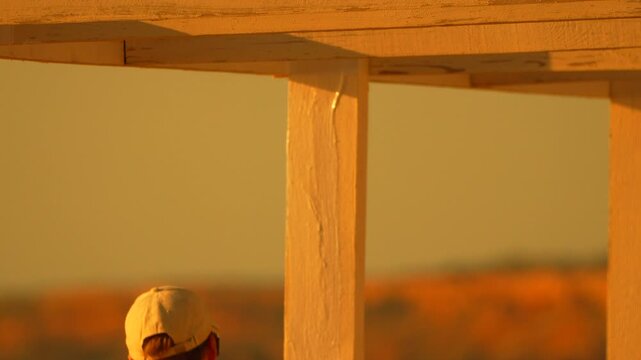 White primer application on wooden gazebo posts by a construction worker during golden hour. Medium tripod shot.