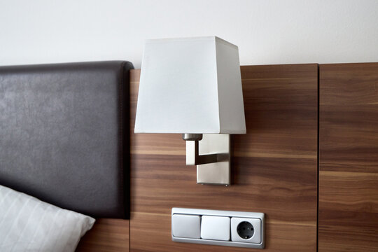 Modern hotel bedside wall lamp with a fabric lampshade, built into a wooden headboard next to a leather pillow, a light switch, and a power socket outlet. 