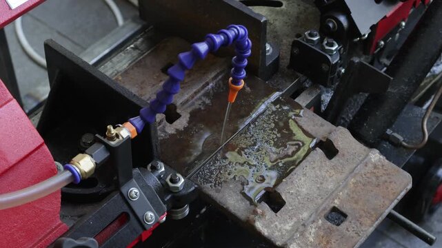 Industrial scene showing a band saw machine in action. A metal part is being precisely cut by a saw blade, while a flexible coolant nozzle delivers lubricant to the cutting zone, reducing friction and