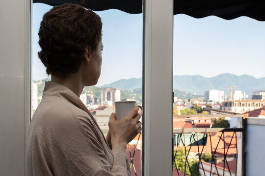 woman after cancer treatment enjoys a warm drink while gazing from an apartment window, portraying quiet self care, emotional wellbeing, recovery and a restorative city stay