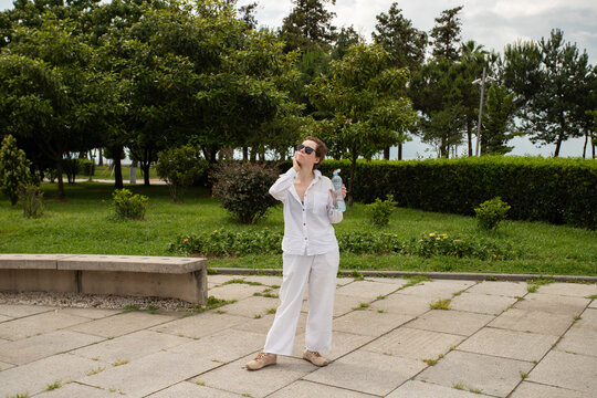woman on a cancer rehabilitation break holds a water bottle and touches her neck outdoors, expressing heat discomfort and self awareness during a hot day