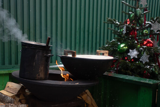 large black cauldrons simmer over open fire with flames and steam rising, rustic outdoor cooking zone decorated with christmas tree and ornaments at a cozy winter festival