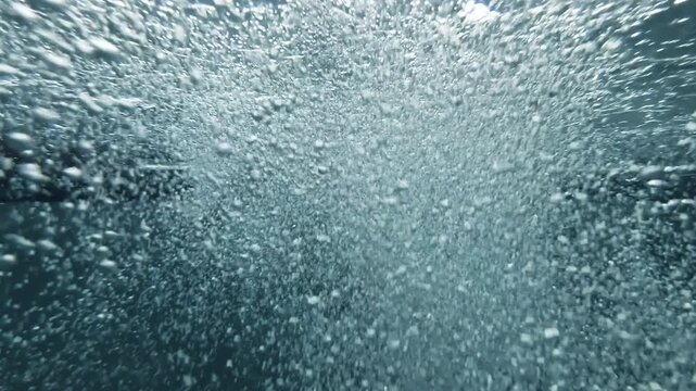 Underwater view of a woman jumping feet first into an indoor swimming pool, creating dramatic bubbles rising toward the surface in slow aquatic motion