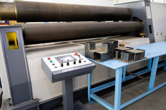 Heavy-duty four-roll bending machine forms metal plates; industrial control panel enables precise operation. Steel profile samples on blue workbench highlight fabrication workflow in a production sett