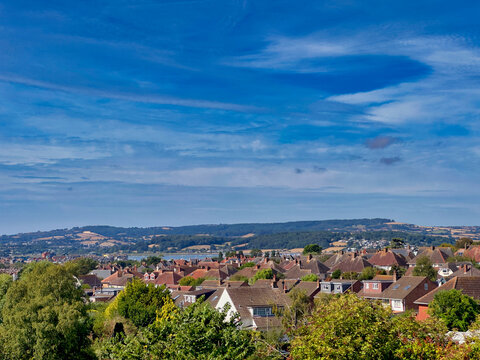 Panoramic view over the coastal town of Exmouth, Devon, featuring residential rooftops, the Exe Estuary, rolling countryside, and a vast blue sky