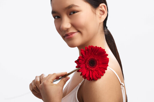 Asian woman holds a red flower near her shoulder, smiling softly against a white studio backdrop, conveying warmth, elegance, and a fresh, serene mood.