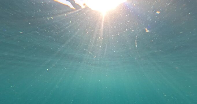 Clear ocean surface transitions into a deep marine view beneath a bright sun. A submerging camera shot captures light rays piercing calm water.