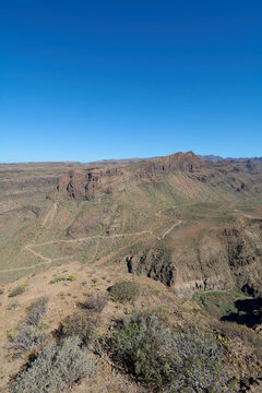 High-angle panoramic view of the majestic volcanic canyons, winding trails, and rugged mountain topography of Degollada las Yeguas under a clear blue sky in Gran Canaria.