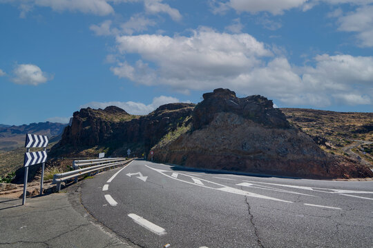 Winding mountain road curve with traffic signs and safety barriers under a cloudy blue sky near Degollada de las Yeguas in Gran Canaria, Spain.