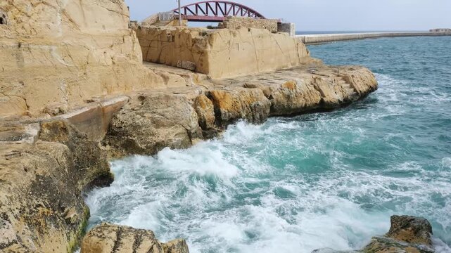 red steel breakwater bridge spans valletta harbour entrance lighthouse distant waves crash rocky foreground shore valletta breakwater st elmo breakwater lighthouse tower grand harbour entrance