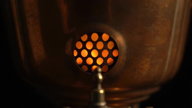 Close-up of a vintage samovar with glowing embers visible through a circular grate. Warm, inviting light emanates from the heating element within.