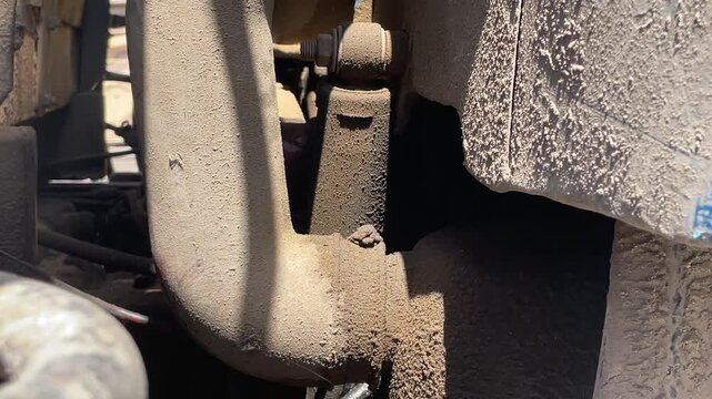 Close view of a dusty truck frame and undercarriage, showing metal brackets, suspension components, wheel parts, dirt, and signs of wear during an open-air mechanical inspection in sunlight.