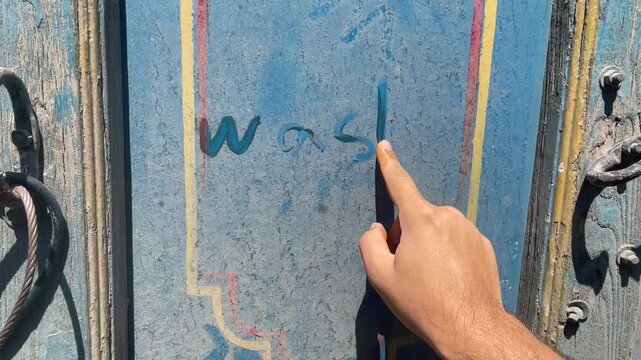 hand points at writing wash me on dusty blue truck body panel, highlighting surface markings and grime during an open-air vehicle inspection, maintenance check, or documentation process at work.