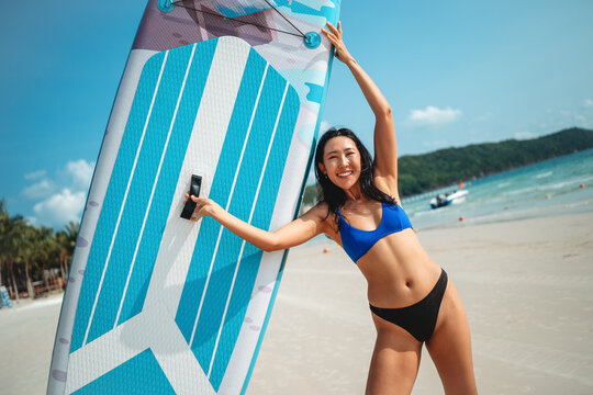 Happy Asian woman posing with a stand-up paddleboard on a sunny tropical beach