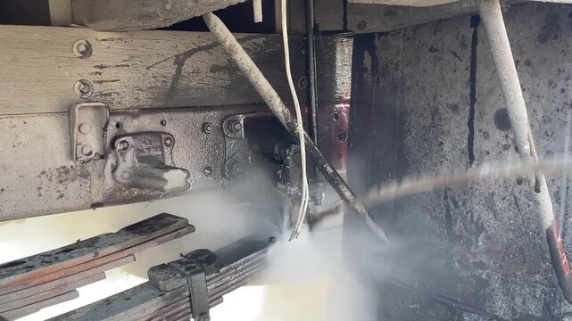 Pressure washing begins beneath a dusty truck, with a strong water jet removing dirt and grime from the undercarriage, suspension, and chassis during professional vehicle cleaning outdoors.