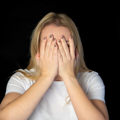 Fototapeta premium Glad smiling blonde female model hiding her face behind hand, has broad smile, happy to recieve compliments from handsome man, isolated against black background. Positive young woman