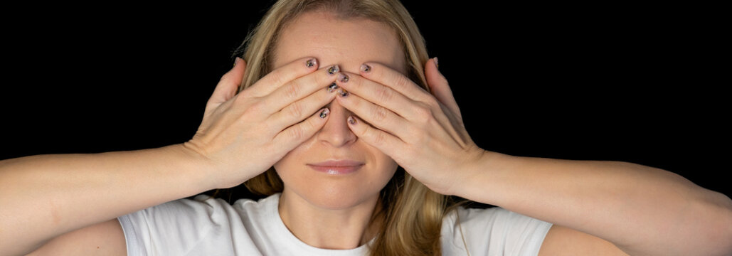 Glad smiling blonde female model hiding her face behind hand, has broad smile, happy to recieve compliments from handsome man, isolated against black background. Positive young woman