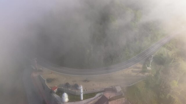 Cinematic 4K footage in gentle slow motion, slowly rising and moving backward above a foggy winding road as drifting mist creates a dramatic and atmospheric coastal landscape.