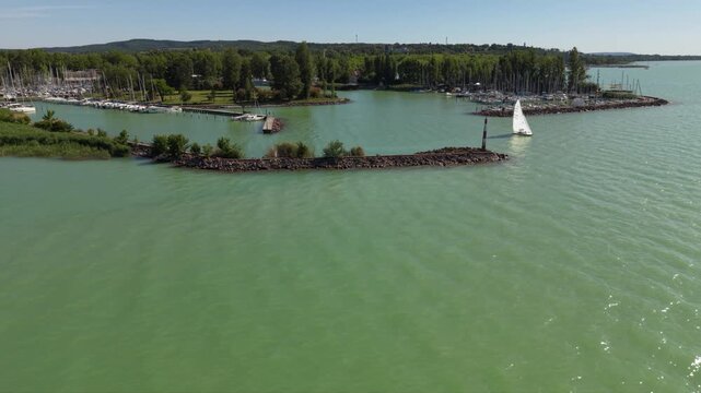 Lake Balaton, Hungary: aerial approach toward a white sailboat passing a marina breakwater, with pale green lake water, moored yachts and a wooded shoreline beneath clear skies.