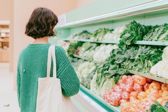 Woman in a green knit sweater seen from behind while browsing fresh leafy vegetables in a modern grocery store for sustainable living concepts
