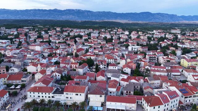 Aerial panoramic view of Novalja, Croatia, showing the coastal town&rsquo;s dense red-roofed architecture, lively central promenade and traditional churches, with the dramatic rocky mountains of Pag Island 