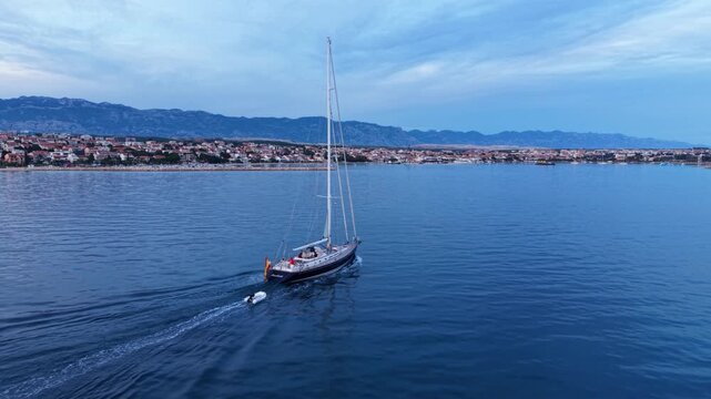 Aerial view of a large sailing yacht cruising across calm Adriatic waters near Novalja, Croatia, with the coastal town and dramatic rocky Velebit mountains in the background.